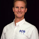 Photo of a white man with short, medium brown hair. He is smiling and in front of a black background. He is wearing a white button down shirt with the initials ACB on it. 