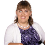 Headshot of a white woman with chin-length brown hair. She is smiling and wearing a purple and black dress, white cardigan and a white bead necklace