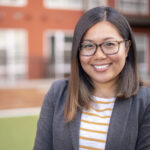 Headshot of an Asian woman with medium length, dark hair, and brown and black glasses. She is smiling and wears a white and yellow striped shirt and a gray jacket. 